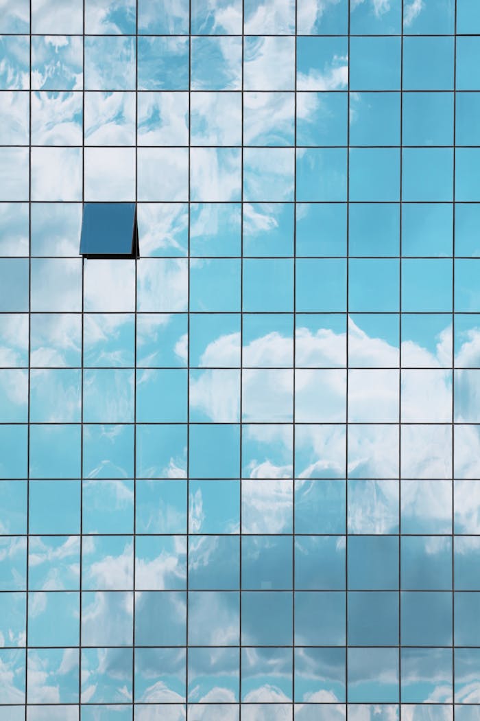 A modern skyscraper facade reflecting a blue sky with clouds and an open window revealing contrast.